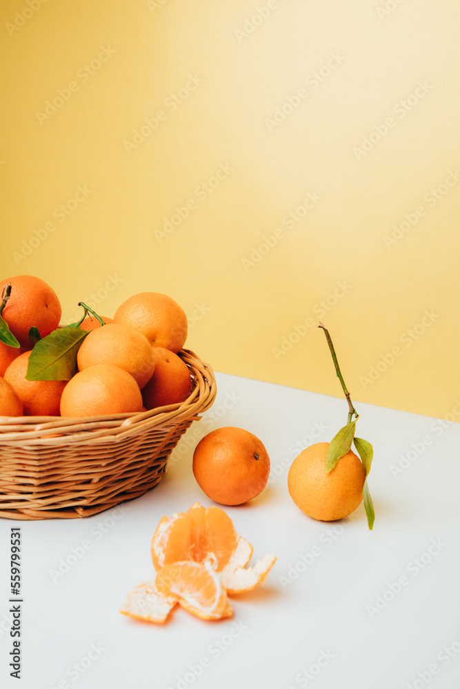Orange mandarins clementine with green leaves in straw basket on a white-yellow background. Front view Orange mandarins clementine with green leaves in straw basket on a white-yellow background. Front view