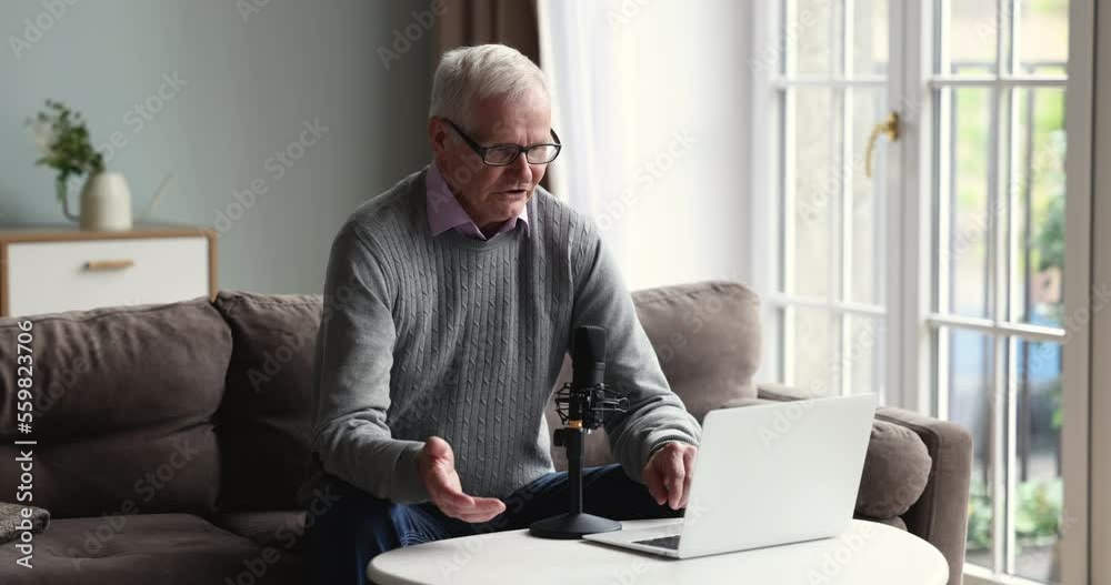 Serious older man in glasses makes speech into mike sit on sofa staring at laptop screen, lead online conversation, communicates with internet audience. Vlogging, streaming, educational webinar event