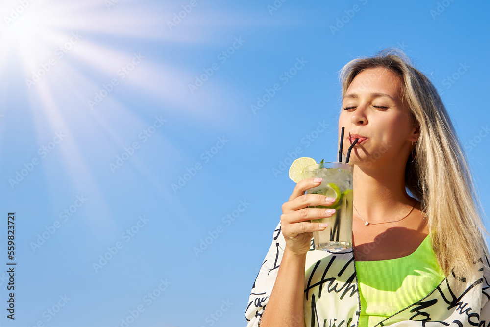 On sunny day, satisfied girl in bright swimsuit enjoys delicious cool cocktail from straw under blue sky. Tanned blonde holds glass of cocktail in her hand, drinks a delicious drink from a straw On sunny day, satisfied girl in bright swimsuit enjoys delicious cool cocktail from straw under blue sky. Tanned blonde holds glass of cocktail in her hand, drinks a delicious drink from a straw