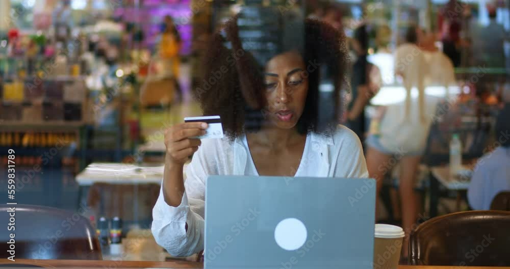 View through cafeteria glass windows of focused multi-ethnic dark-skinned woman, holding credit card, transferring data to laptop, making online cashless payment via mobile app or internet banking View through cafeteria glass windows of focused multi-ethnic dark-skinned woman, holding credit card, transferring data to laptop, making online cashless payment via mobile app or internet banking