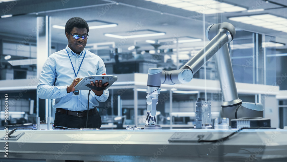Young Black Male Engineer Using a Tablet Computer, Researching and Developing a Futuristic Robotic Arm Machine in a High Tech Industrial Laboratory with Modern Equipment.