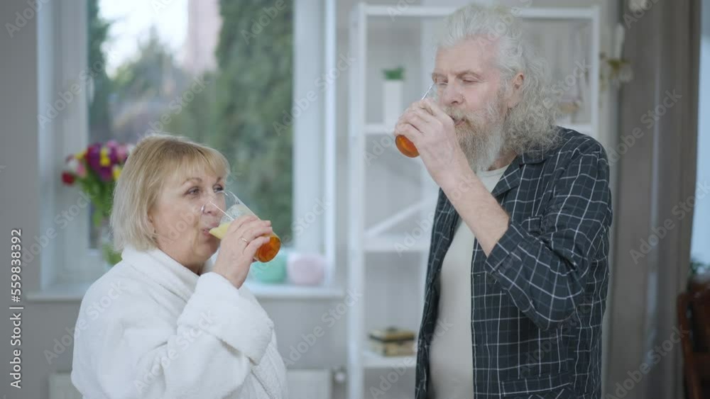 Senior positive couple drinking healthful tasty banana smoothie standing at home indoors. Happy confident Caucasian husband and wife enjoying delicious healthy drink in the morning smiling Senior positive couple drinking healthful tasty banana smoothie standing at home indoors. Happy confident Caucasian husband and wife enjoying delicious healthy drink in the morning smiling