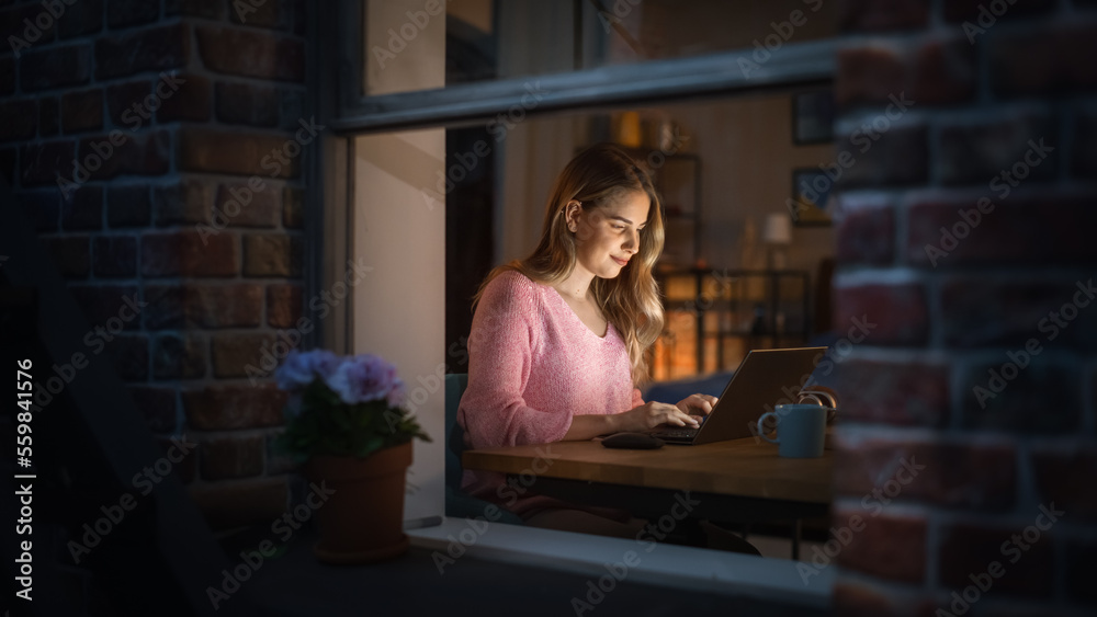 Portrait Shot of a Beautiful Smiling Young Woman Working on a Laptop at Her Desk in the Cozy Bedroom. Happy Female Thinking and Chatting on Social Media. View Through the Window From Outside Portrait Shot of a Beautiful Smiling Young Woman Working on a Laptop at Her Desk in the Cozy Bedroom. Happy Female Thinking and Chatting on Social Media. View Through the Window From Outside