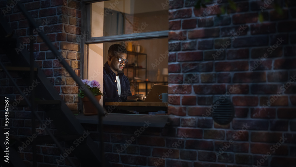 Portrait of Stylish Black Businessman Works on Laptop, Does Data Analysis While Working from Home at Night. Young Male Digital Entrepreneur Works on e-Commerce Startup Project. View Through the Window