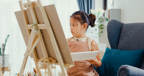 Asian toddler girl with apron sit on sofa chair in front of painting canvas with color palette use paint brush focus on painting abstract on weekend at home. Creative lifestyle for kid concept.