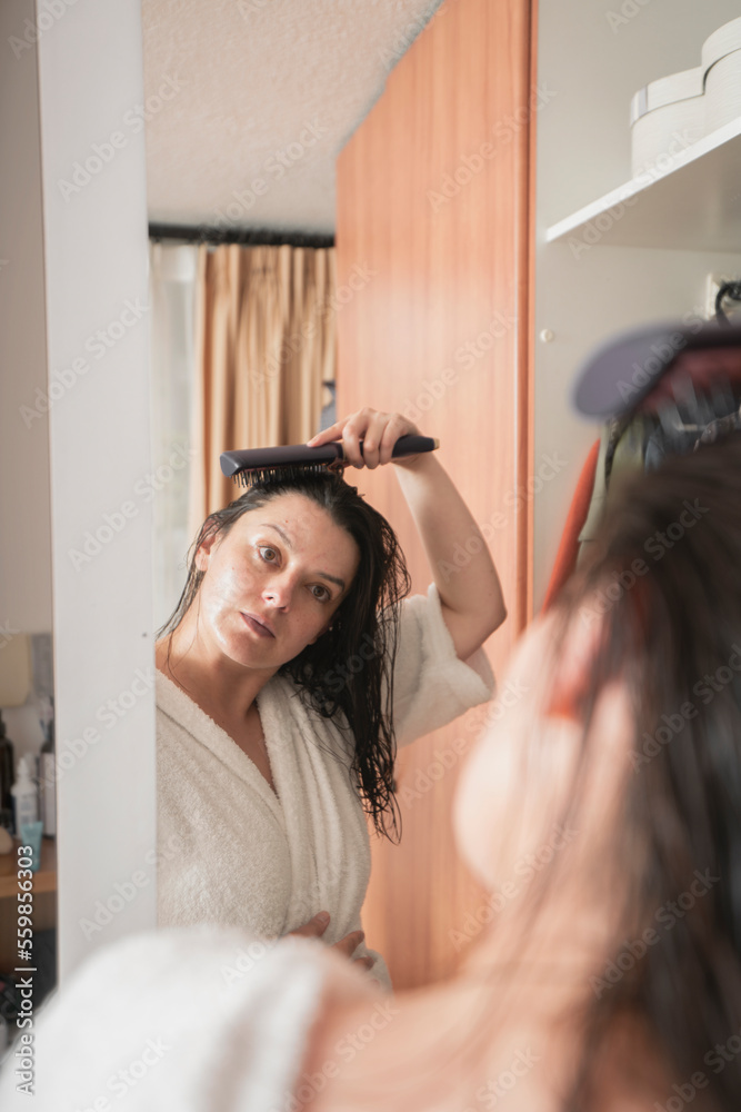 Fototapeta premium woman in a white bathrobe combing her hair with a brush in front of the mirror on her closet door in her bedroom