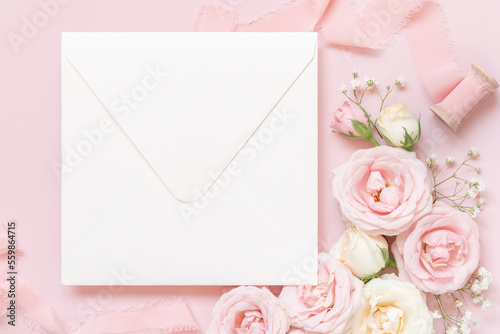 Blank square envelope between pink roses and pink silk ribbons on pink top view, wedding mockup