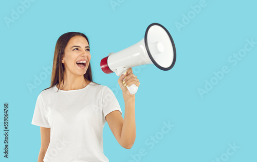 Smiling young woman shouting into megaphone making recommendation. Attractive excited brunette girl announcing with loudspeaker on light blue background, copy space