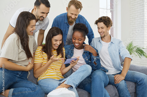 Group of people gathered together around female friend with mobile phone and are watching funny photos and videos together. Smiling multiracial millennial friends relaxing together on comfortable sofa