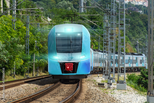 Suburban electric train of blue color goes turn from a distance with the headlights on along the way among the green trees.