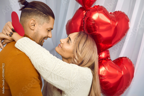 Young brunette man and blonde woman holding heart in her hands looking at each other and hugging with red foil ballons background. Happy couple in love. Care, tenderness and Valentines day concept.