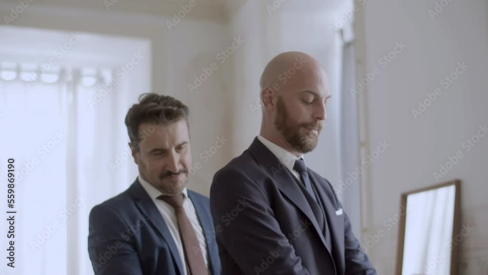 Handsome gay helping partner putting on his jacket. Cheerful Caucasian homosexual couple in official suits getting ready for wedding ceremony, laughing and kissing. LGBT, marriage, love concept