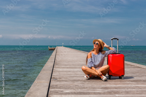 A lady is sitting on a pier against the background of the sea with a red suitcase for luggage, shorts, sunglasses, travel 2023, summer mood, open borders