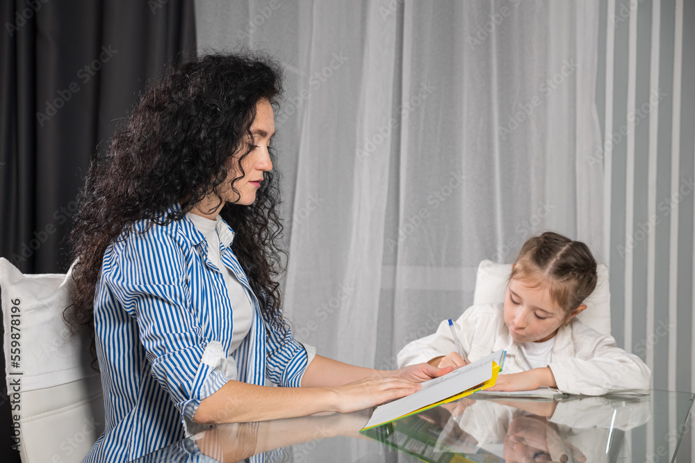 Nervous mother helps daughter with doing homework in apartment and explains material. Excited junior schoolgirl writes in notebook and listen to mom at table Nervous mother helps daughter with doing homework in apartment and explains material. Excited junior schoolgirl writes in notebook and listen to mom at table