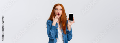 Impressed and astonished redhead woman seeing something unbelievable, spilling tea in internet, gasping cover opened mouth and showing samrtphone display, white background photo