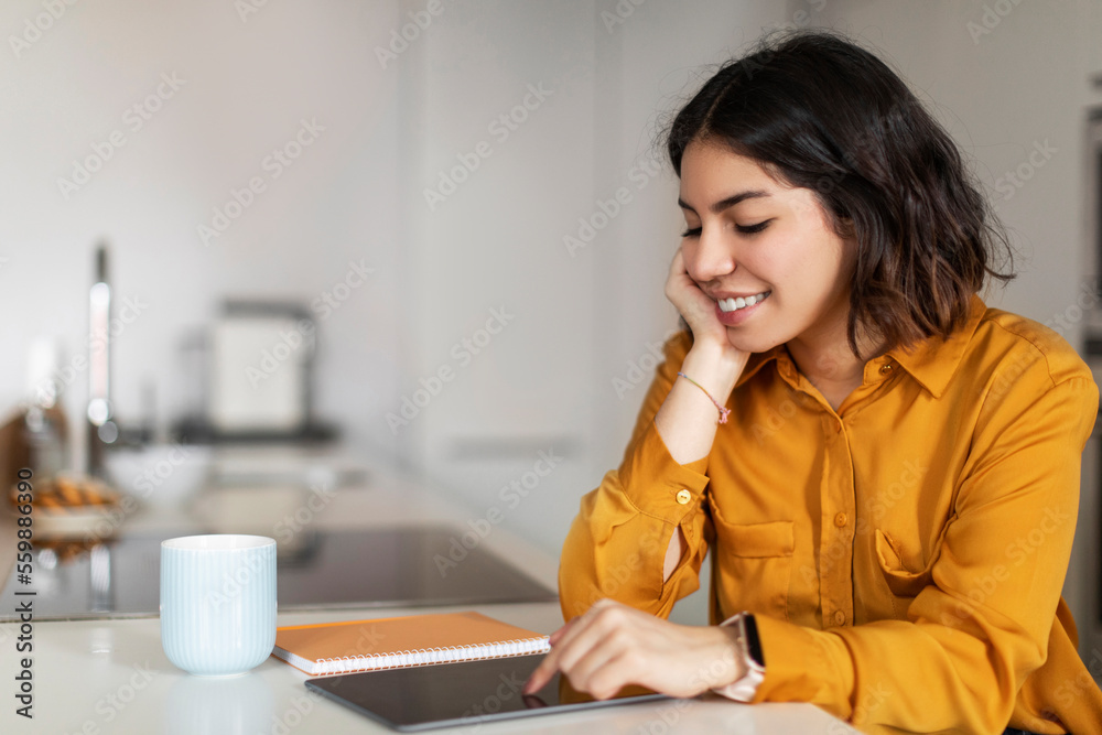 Arab Woman Browsing New App On Digital Tablet While Relaxing In Kitchen Arab Woman Browsing New App On Digital Tablet While Relaxing In Kitchen