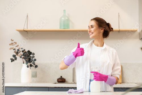 Attractive housewife holds a spray bottle with cleaning agent in her hand and shows a thumbs up. 
