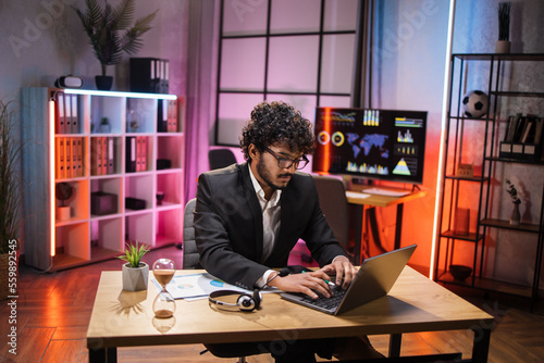 Side view, portrait of young smiling experienced smart stylish bearded manager in eyeglass, businessman in suit sitting at table, using laptop in evening modern office