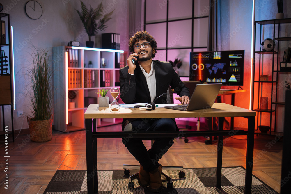 Full length portrait of handsome concentrated hardworking bearded businessman or manager in suit having business conversation with smart phone and laptop computer in evening office.