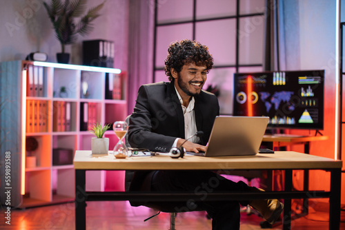 Portrait of handsome experienced smart stylish curly bearded manager, businessman, wearing suit sitting at table, using laptop at evening time in modern office.