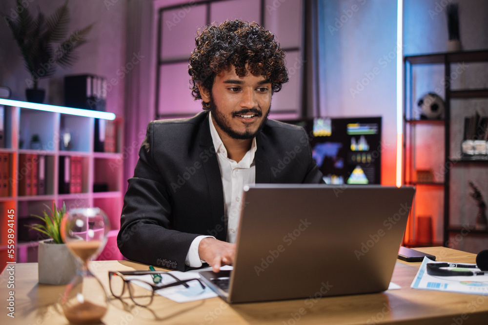 Handsome experienced smart stylish curly bearded manager, broker, businessman, wearing suit sitting at table, using laptop at evening time in modern office. Handsome experienced smart stylish curly bearded manager, broker, businessman, wearing suit sitting at table, using laptop at evening time in modern office.