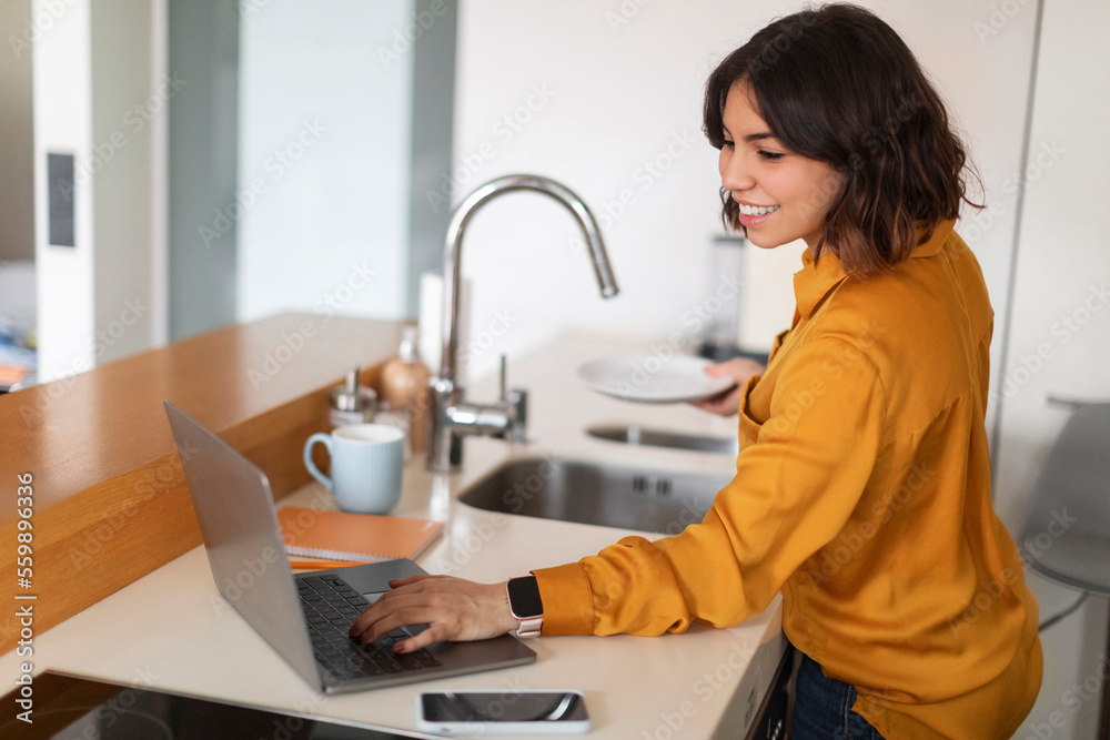 Young Arab Female Using Laptop Computer And Washing Dishes In Kitchen Young Arab Female Using Laptop Computer And Washing Dishes In Kitchen