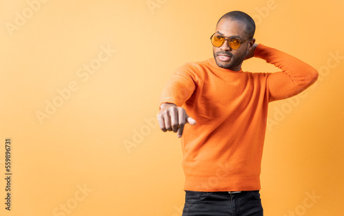A dark-skinned young man dances in front of an orange street wall, the man is wearing a jumper and orange sunglasses.