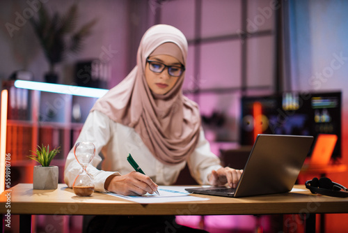 Focus in hand with pen. Attractive confident muslim business woman, office manager, wearing hijab using laptop while making financial report while writing on paper working at night.