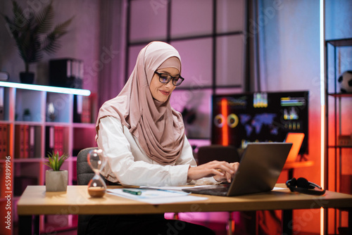 Experienced muslim businesswoman, economist, broker in hijab wearing eyeglass sitting at table with laptop at evening time at office. Concept of deadline of project.