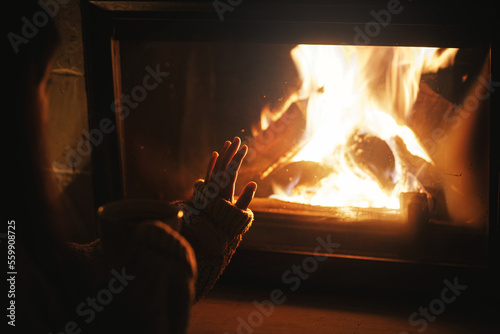 Woman holding cup of tea and warming up hands at cozy fireplace in dark evening room. Fireplace heating in house, electricity blackout. Atmospheric time at fireside in home