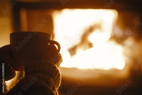 Woman holding cup of tea and warming up hands at cozy fireplace in dark evening room, close up. Fireplace heating in house, electricity blackout. Atmospheric time at fireside in home