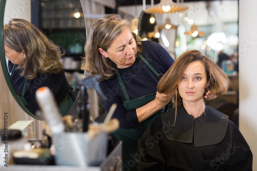 Elderly woman professional hair stylist discussing young female customer preferences in salon, choosing new hairdo .