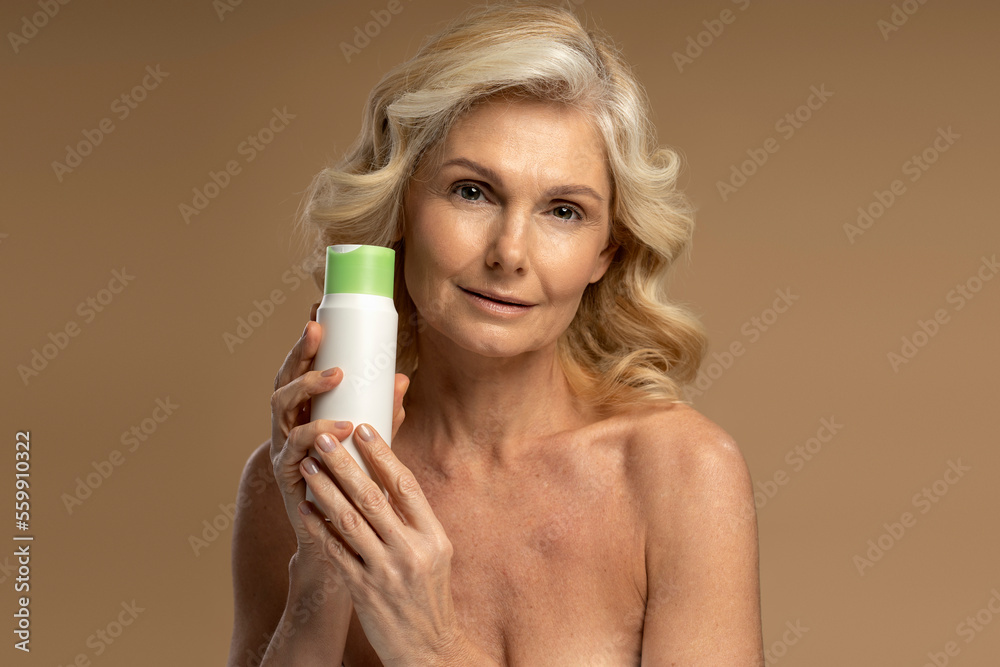 Confident beautiful Caucasian shirtless, 50s mature woman looking at camera, holding a jar with anti-aging smoothing moisturizing cosmetic product, isolated beige background. Mature beauty concept Confident beautiful Caucasian shirtless, 50s mature woman looking at camera, holding a jar with anti-aging smoothing moisturizing cosmetic product, isolated beige background. Mature beauty concept