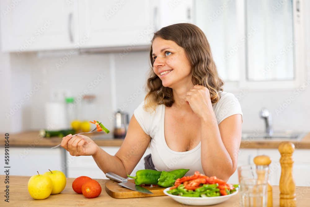 Portrait of cheerful european woman leaning on table in kitchen and eating just cooked salad. Woman enjoying meal.