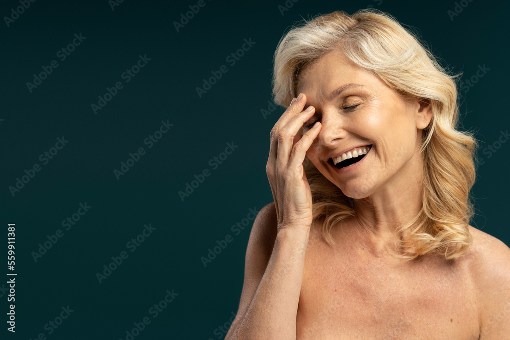 Caucasian attractive blonde mature woman with her eyes closed, smiling a beautiful toothy smile, touching her face, laughing, isolated over dark colored background. Skin care concept Caucasian attractive blonde mature woman with her eyes closed, smiling a beautiful toothy smile, touching her face, laughing, isolated over dark colored background. Skin care concept