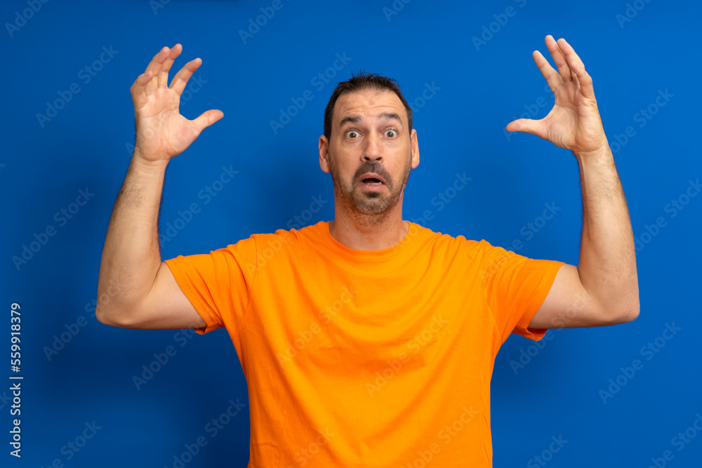 Hispanic man dreamy bearded melancholic surprised attractive man 40s in casual orange basic t-shirt spreading hands isolated on blue color background studio portrait. People lifestyle concept. Hispanic man dreamy bearded melancholic surprised attractive man 40s in casual orange basic t-shirt spreading hands isolated on blue color background studio portrait. People lifestyle concept.