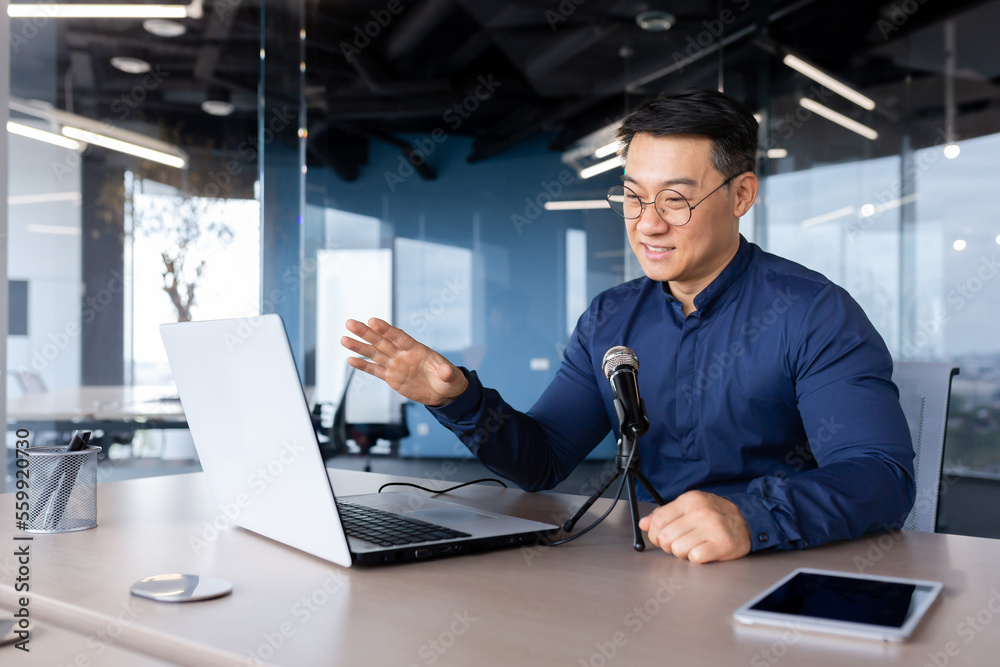 A young Asian man is sitting at a desk in the office. Speaks into the microphone through a video call from a laptop. Online business meeting, blogger, recording interviews, podcast. A young Asian man is sitting at a desk in the office. Speaks into the microphone through a video call from a laptop. Online business meeting, blogger, recording interviews, podcast.