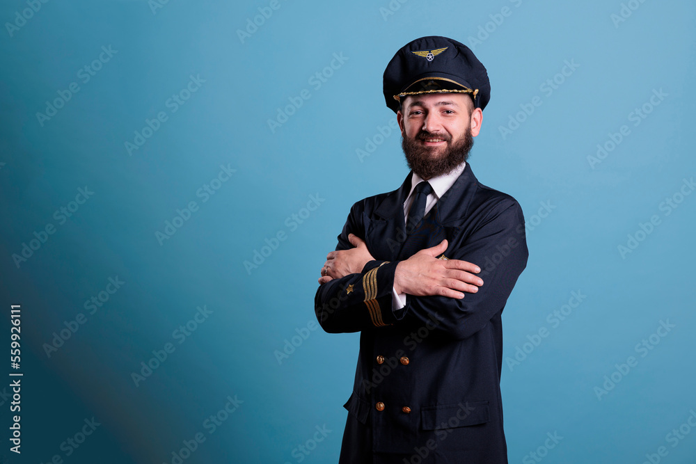 Airplane pilot wearing uniform and hat portrait, confident plane captain standing with crossed arms, looking at camera. Smiling civil aviator with badge on professional suit Airplane pilot wearing uniform and hat portrait, confident plane captain standing with crossed arms, looking at camera. Smiling civil aviator with badge on professional suit