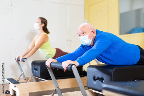 Focused senior man wearing medical mask to protect against viral infections practicing pilates stretching exercises on reformer as part of remedial gymnastics.. photo