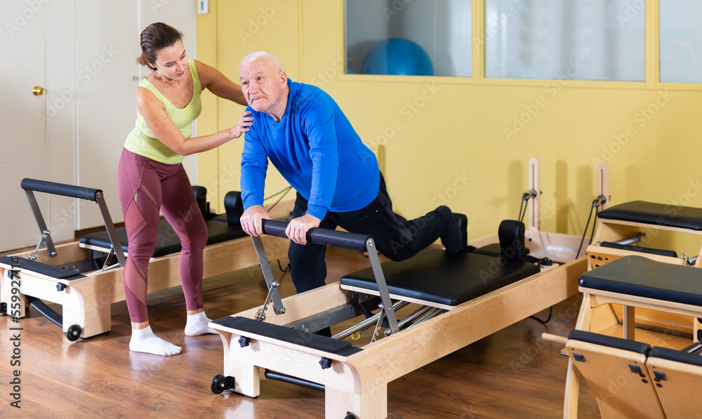 Personal female trainer controlling movements of senior man doing pilates on reformer in fitness studio. Healthy active lifestyle
