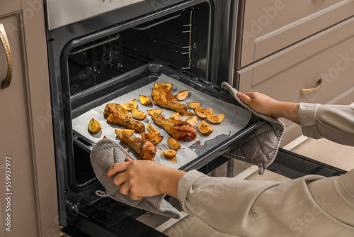 Woman taking baking tray with paper, chicken legs and potato from oven, closeup
