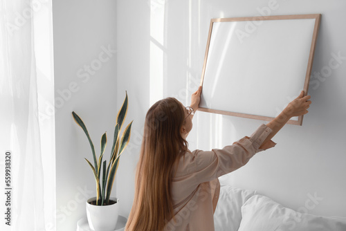 Young woman hanging blank frame on light wall in bedroom