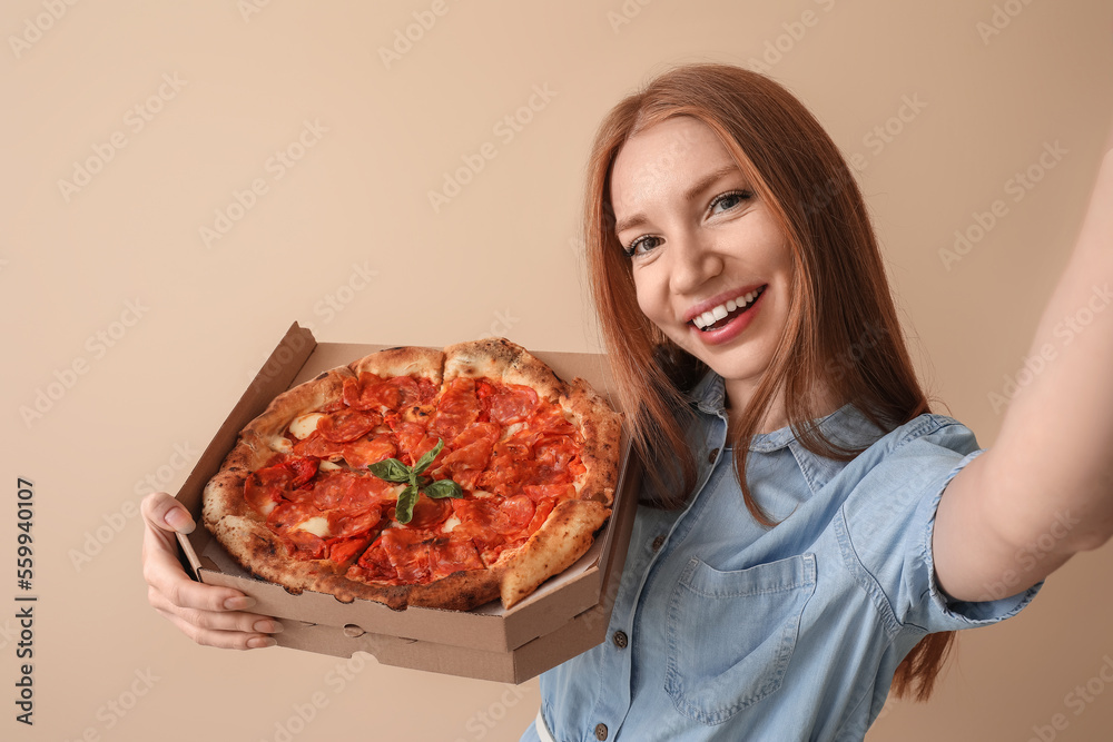 Young woman with box of tasty pizza taking selfie on beige background