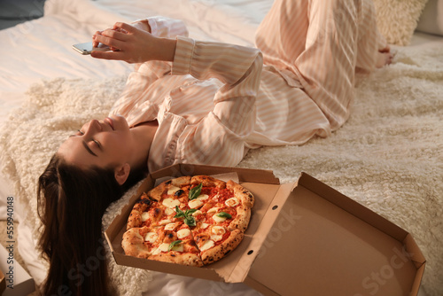 Young woman with tasty pizza using mobile phone in bedroom at night
