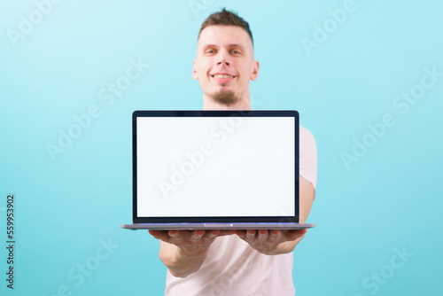 A man holding a laptop with two hands and presenting something on an empty blank screen and looking at the camera on blue background. Present. Performance. Studio Shot. Modern. Attractive. Commercial