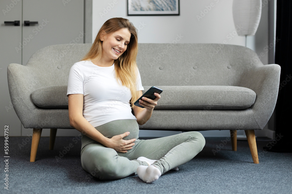 Young beautiful pregnant woman holding mobile phone touching belly shopping online sitting in floor at home. Smiling future mother using mobile app. Pregnancy concept 