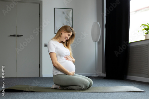 Happy pregnant woman sitting on the floor and touching her belly at home. Pregnancy, people and recreation concept 