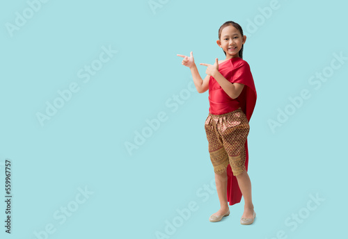 Full body Asian little girl in Thai national costumes with pointing finger blank workspace, isolated on pastel plain light blue background