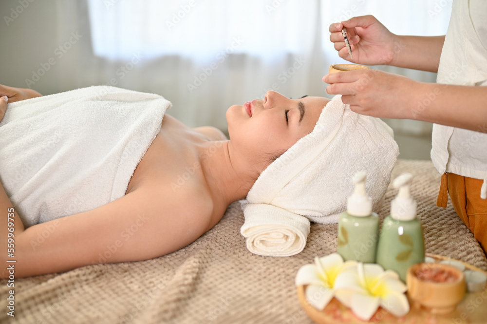 Asian woman lying on massage table, receiving facial treatment clay mask by a spa worker. Asian woman lying on massage table, receiving facial treatment clay mask by a spa worker.