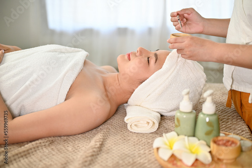 Asian woman lying on massage table, receiving facial treatment clay mask by a spa worker.